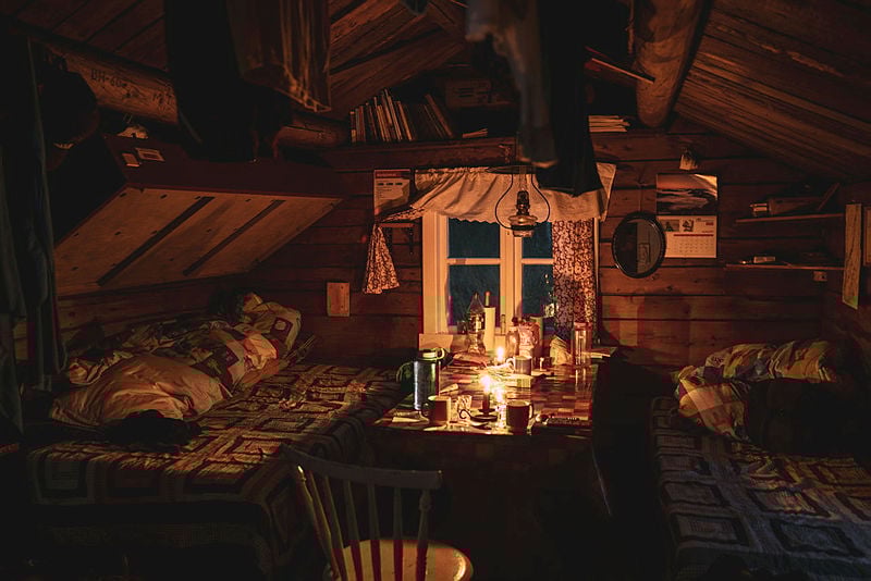Ei hyttestue med tre senger og et bord imellom. Et hvitkarmet vindu. En stol i forgrunnen. Det står mange ting på bordet. Det er mørkt ute og et lys på bordet.